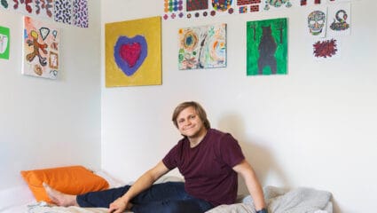 En ungdom sitter i en säng under en mängd konstverk och mandalas