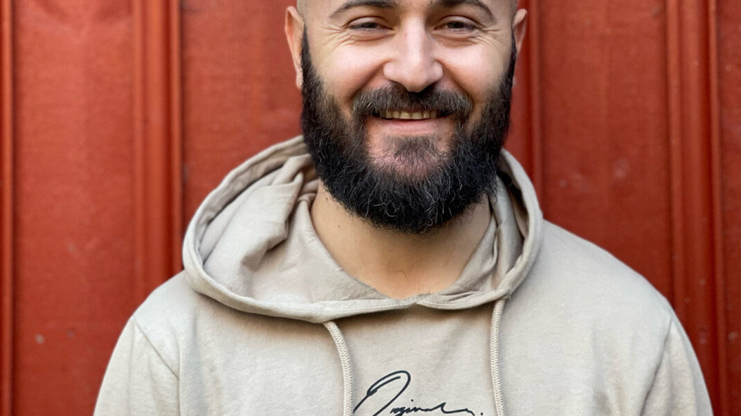 Porträtt på Ervis Kabashi i beige Hood-tröja mot röd trävägg