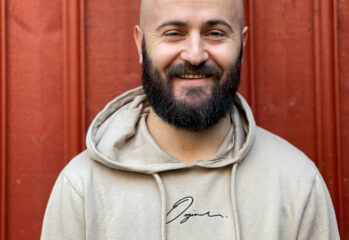 Porträtt på Ervis Kabashi i beige Hood-tröja mot röd trävägg