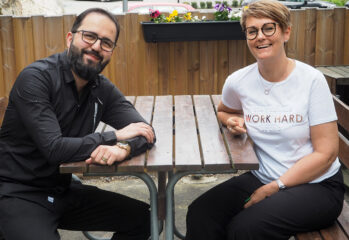 Bilal i svart tröja och Petra i vit t-shirt sitter vid ett bord, tittar in i kameran