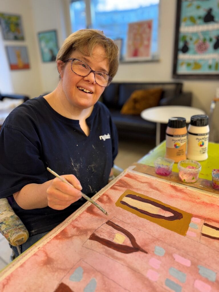 En kvinna sitter i en ateljé och målar på en färgstark tavla med geometriska former i rosa, brunt och gult. Hon ler mot kameran och håller en pensel i handen. Bakom henne syns fler tavlor på väggarna och ett fönster som släpper in dagsljus.