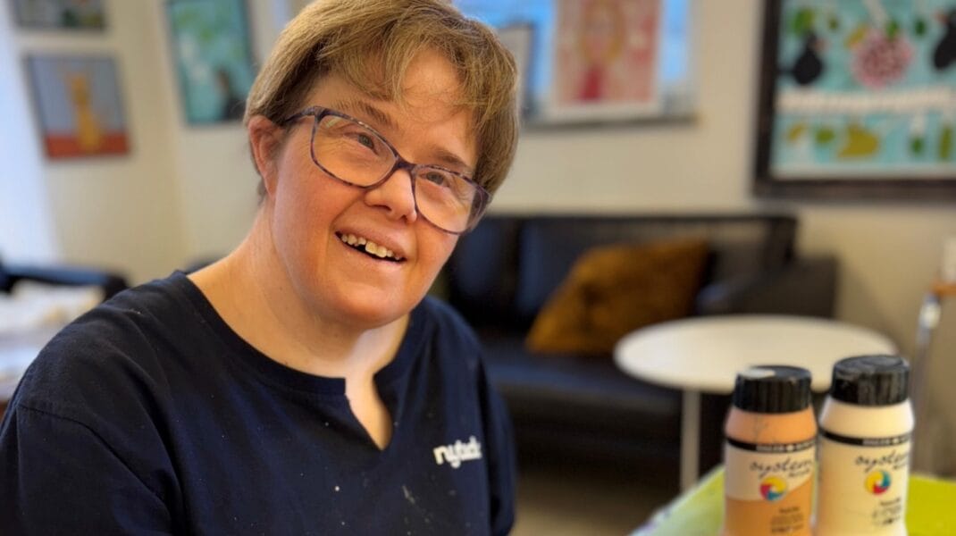 En kvinna sitter i en ateljé och målar på en färgstark tavla med geometriska former i rosa, brunt och gult. Hon ler mot kameran och håller en pensel i handen. Bakom henne syns fler tavlor på väggarna och ett fönster som släpper in dagsljus.