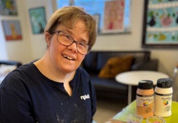 En kvinna sitter i en ateljé och målar på en färgstark tavla med geometriska former i rosa, brunt och gult. Hon ler mot kameran och håller en pensel i handen. Bakom henne syns fler tavlor på väggarna och ett fönster som släpper in dagsljus.