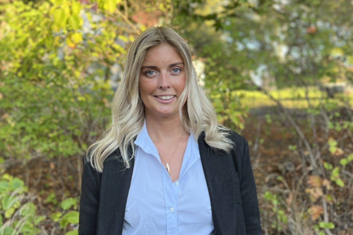 Porträtt av en leende kvinna med blont hår, klädd i ljusblå skjorta och mörk kavaj, fotograferad utomhus i grönskande miljö.