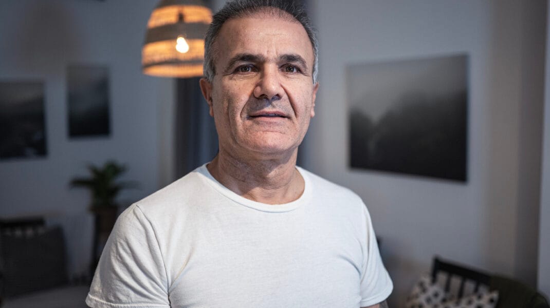 En person står inomhus i ett möblerat rum och tittar in i kameran. Personen bär en vit T-shirt och bakgrunden visar stämningsfull belysning, väggkonst och sittmöbler.