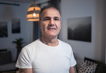 En person står inomhus i ett möblerat rum och tittar in i kameran. Personen bär en vit T-shirt och bakgrunden visar stämningsfull belysning, väggkonst och sittmöbler.