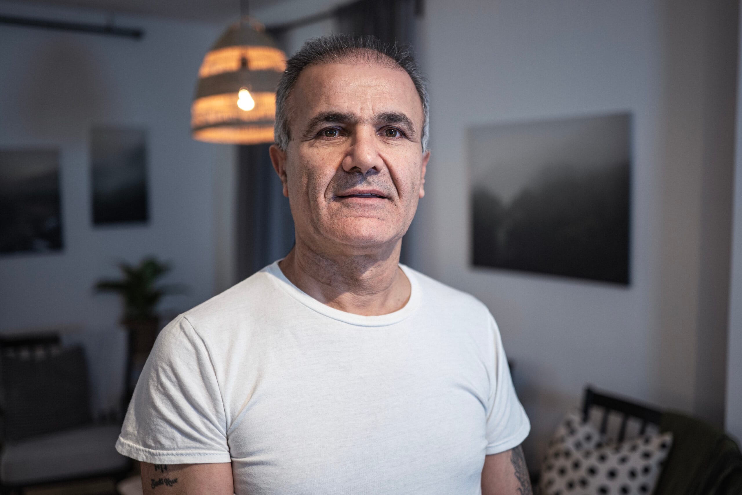 ”En person står inomhus i ett möblerat rum och tittar in i kameran. Personen bär en vit T-shirt och bakgrunden visar stämningsfull belysning, väggkonst och sittmöbler.