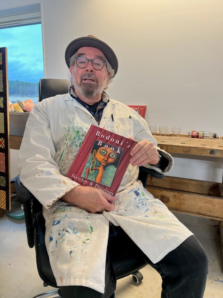 En person sitter i en ateljé och håller upp en bok med illustrationer, iklädd arbetsrock med färgfläckar, med konstmaterial i bakgrunden.