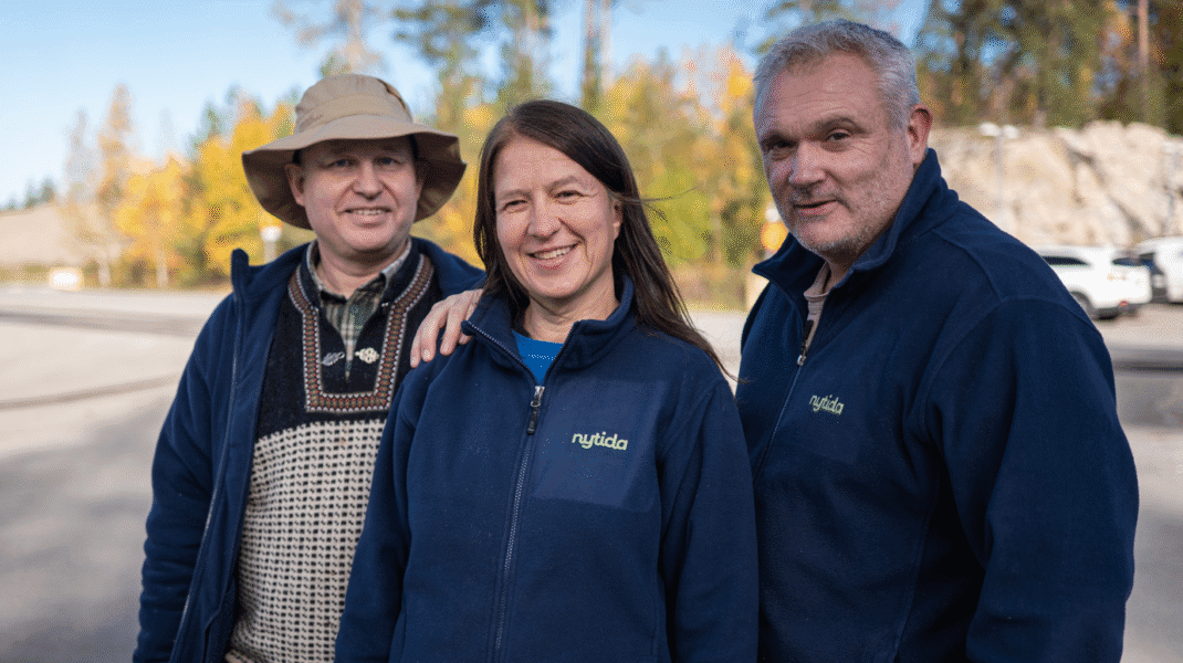 Tre personer står utomhus nära varandra och ler mot kameran. De bär mörkblå jackor med logotyp och har träd och höstfärger i bakgrunden.