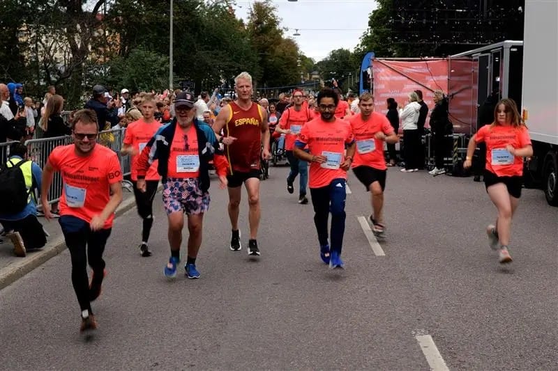 En grupp personer springer tillsammans på en avstängd gata under ett motionslopp. De bär matchande tröjor med startnummer och publik står längs sidorna.