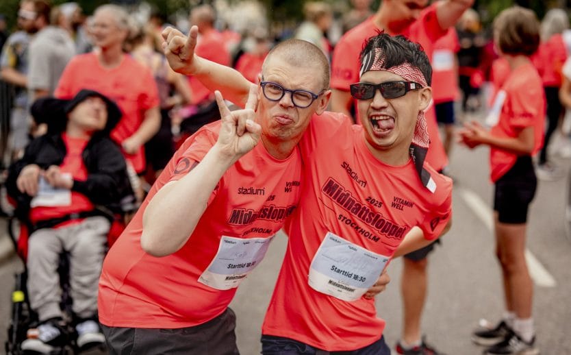 Två personer som ler mot kameran med människor i bakgrunden. Båda personerna är iklädda röda, kortärmade sporttröjor med texten ”Midnattsloppet” tryckt på bröstet. En av personerna bär solglasögon och den andra vanliga glasögon.
