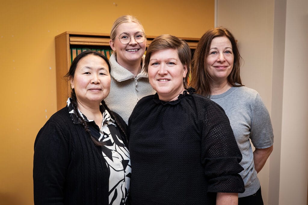 Fyra personer står inomhus tätt tillsammans och ler mot kameran. De befinner sig i ett rum med en bokhylla i bakgrunden.