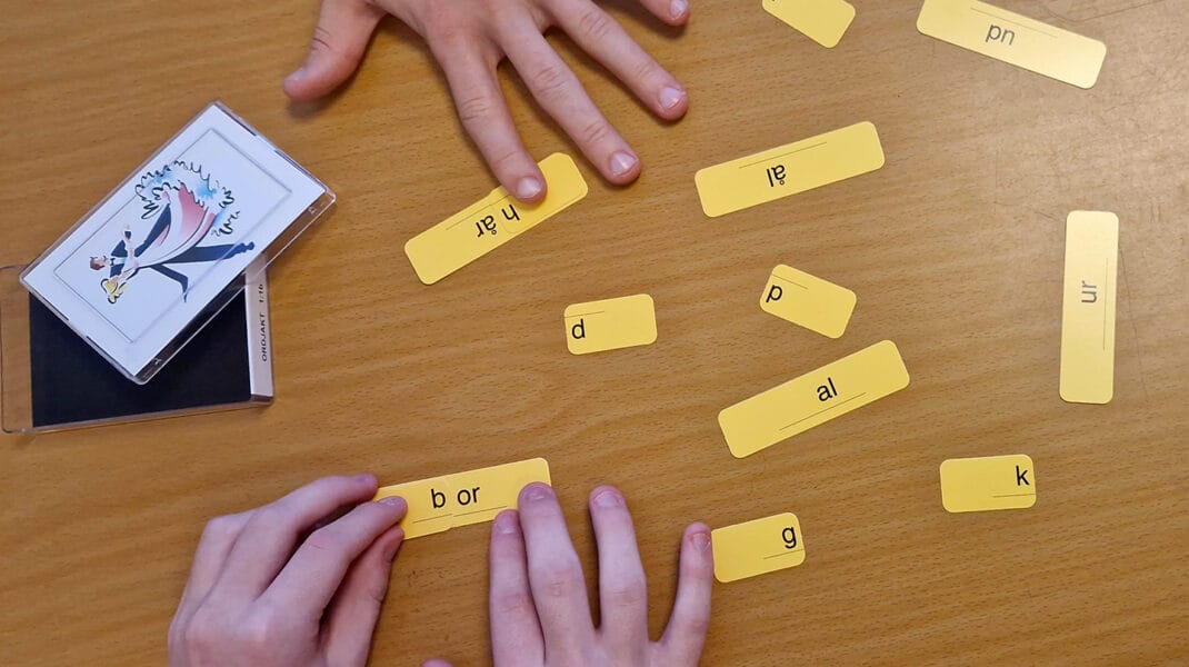 Två personers händer ligger på ett bord där gula kort med bokstäver och ljud kombineras till ord, som en del av en språkövning. Ett kortspel med bildkort ligger bredvid.