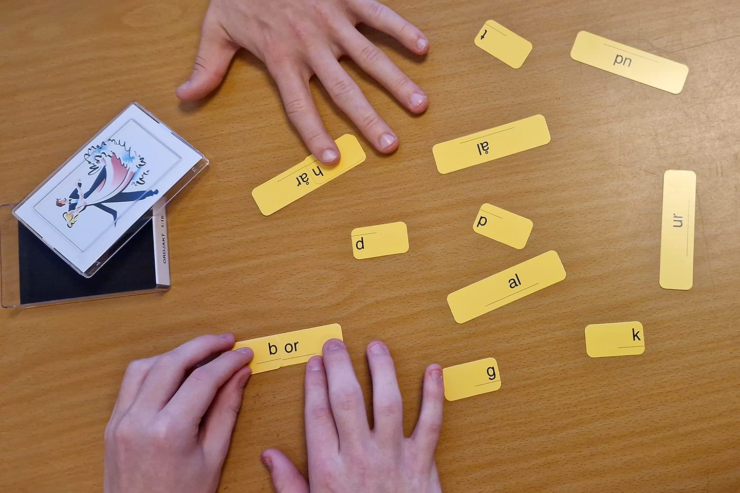 Två personers händer ligger på ett bord där gula kort med bokstäver och ljud kombineras till ord, som en del av en språkövning. Ett kortspel med bildkort ligger bredvid.