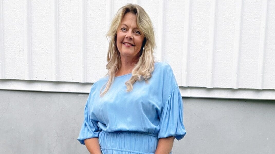 Kvinna med blont, axellångt hår står framför en ljus träfasad och ler mot kameran. Hon bär en ljusblå klänning med korta ärmar och en klocka på handleden.