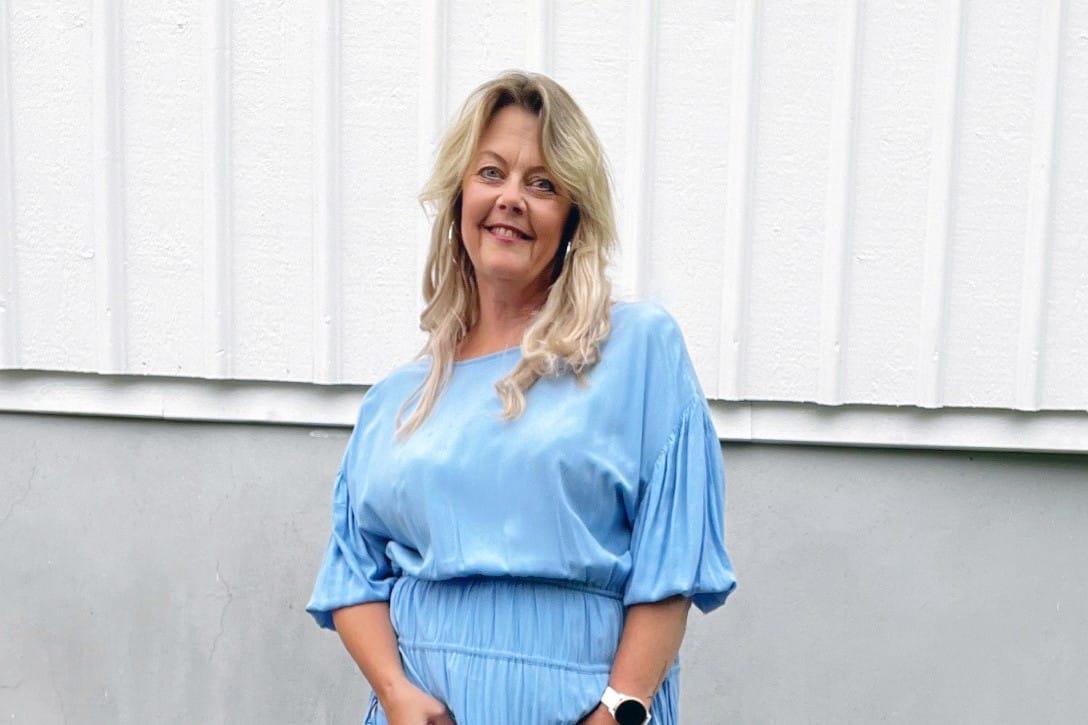 Kvinna med blont, axellångt hår står framför en ljus träfasad och ler mot kameran. Hon bär en ljusblå klänning med korta ärmar och en klocka på handleden.