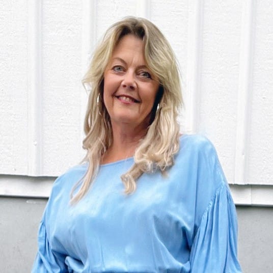 Kvinna med blont, axellångt hår står framför en ljus träfasad och ler mot kameran. Hon bär en ljusblå klänning med korta ärmar.