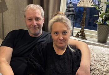 Ett par sitter tätt tillsammans i en soffa hemma. Mannen har armen runt kvinnan och båda tittar mot kameran. Bakom dem syns ett fönster, en bordslampa och en grön växt som skapar en varm och hemtrevlig miljö.