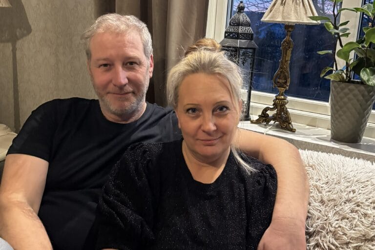 Ett par sitter tätt tillsammans i en soffa hemma. Mannen har armen runt kvinnan och båda tittar mot kameran. Bakom dem syns ett fönster, en bordslampa och en grön växt som skapar en varm och hemtrevlig miljö.