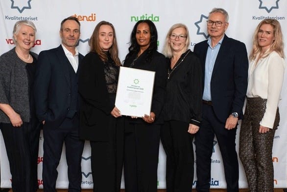 En grupp på sju personer står uppställda framför en bakgrund med logotyper och håller ett diplom från Kvalitetspriset. De är klädda i formella kläder och poserar tillsammans, där en person i mitten håller upp utmärkelsen.