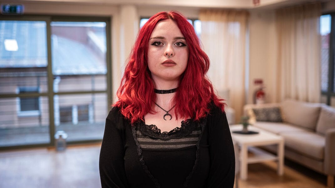 En ung person med rött axellångt hår står inomhus i ett ljust rum med soffa och fönster i bakgrunden.