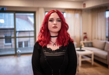 En ung person med rött axellångt hår står inomhus i ett ljust rum med soffa och fönster i bakgrunden.