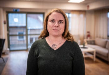 Porträtt av en kvinna med axellångt ljusbrunt hår som står i ett ljust vardagsrum med soffa och stora fönster i bakgrunden.