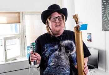 En person med glasögon och svart hatt står inomhus och håller upp en pokal i ena handen och ett inslaget paket i den andra. Personen bär en svart t-shirt med vargmotiv och står i ett ljust rum med fönster och möbler i bakgrunden.