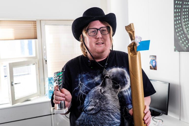 En person med glasögon och svart hatt står inomhus och håller upp en pokal i ena handen och ett inslaget paket i den andra. Personen bär en svart t-shirt med vargmotiv och står i ett ljust rum med fönster och möbler i bakgrunden.