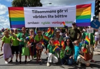 En grupp människor deltar i en prideparad och håller upp en stor regnbågsfärgad banderoll med texten ”Tillsammans gör vi världen lite bättre”. Flera i gruppen bär gröna tröjor och viftar med regnbågsflaggor.