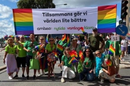 En grupp människor deltar i en prideparad och håller upp en stor regnbågsfärgad banderoll med texten ”Tillsammans gör vi världen lite bättre”. Flera i gruppen bär gröna tröjor och viftar med regnbågsflaggor.