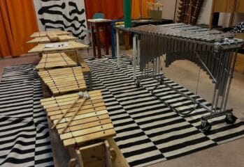 Ett musikrum med flera slagverksinstrument uppställda på en svartvit randig matta. I förgrunden ligger flera träxylofoner med klubbor ovanpå, och längre bak står en större vibrafon på hjul. I bakgrunden syns färgglada väggar och ytterligare instrument.