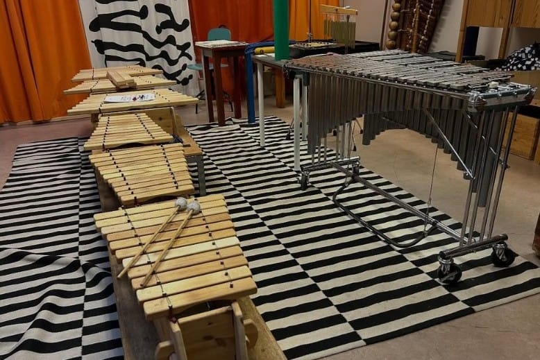 Ett musikrum med flera slagverksinstrument uppställda på en svartvit randig matta. I förgrunden ligger flera träxylofoner med klubbor ovanpå, och längre bak står en större vibrafon på hjul. I bakgrunden syns färgglada väggar och ytterligare instrument.