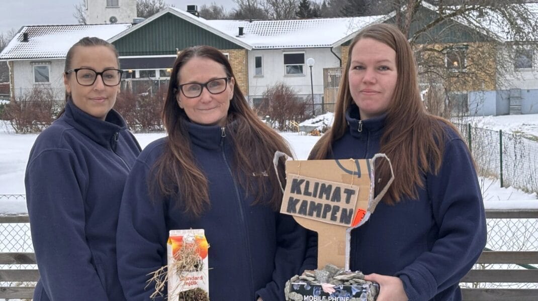 Tre kvinnor står utomhus i vintermiljö framför en byggnad med snötäckt tak. De bär mörkblå fleecejackor och tittar mot kameran. Två av dem håller upp föremål: en hemgjord pokal i kartong med texten “Klimatkampen” samt en dekorerad förpackning med naturmaterial. I bakgrunden syns träd, staket och bostadshus i ett snöigt område.
