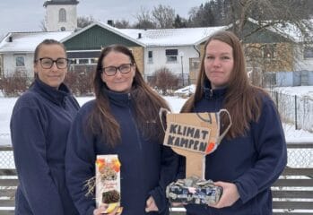 Tre kvinnor står utomhus i vintermiljö framför en byggnad med snötäckt tak. De bär mörkblå fleecejackor och tittar mot kameran. Två av dem håller upp föremål: en hemgjord pokal i kartong med texten “Klimatkampen” samt en dekorerad förpackning med naturmaterial. I bakgrunden syns träd, staket och bostadshus i ett snöigt område.