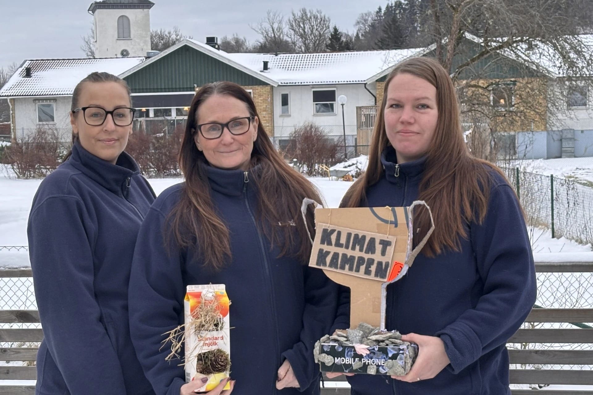Tre kvinnor står utomhus i vintermiljö framför en byggnad med snötäckt tak. De bär mörkblå fleecejackor och tittar mot kameran. Två av dem håller upp föremål: en hemgjord pokal i kartong med texten “Klimatkampen” samt en dekorerad förpackning med naturmaterial. I bakgrunden syns träd, staket och bostadshus i ett snöigt område.