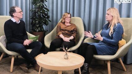 Tre personer sitter i fåtöljer i en samtalsmiljö och deltar i en intervju eller paneldiskussion. En person till vänster håller i ett kort, medan personen till höger gestikulerar under samtalet. I bakgrunden syns gardiner, en växt och en liten rund bordsyta med en vas. Logotypen “Nytida” syns i övre högra hörnet.