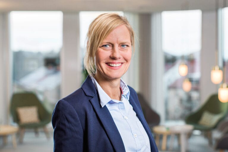 Porträtt av en leende kvinna i kavaj och skjorta som står i en ljus, modern inomhusmiljö med stora fönster och sittgrupper i bakgrunden.