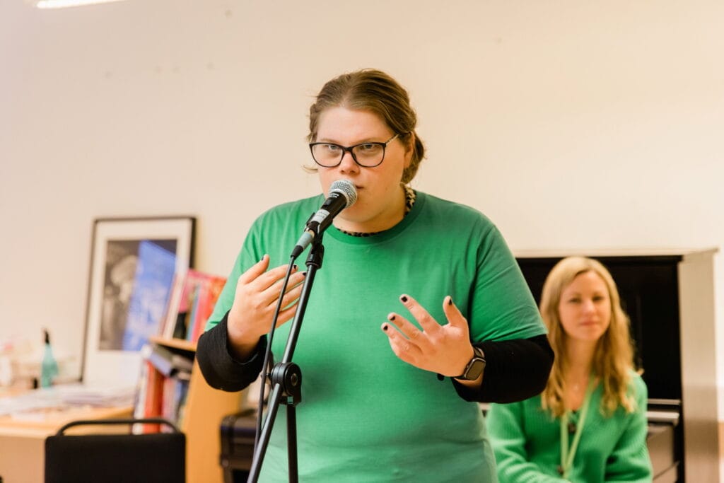 Person i grön tröja talar i mikrofon och gestikulerar, med en sittande person i bakgrunden.