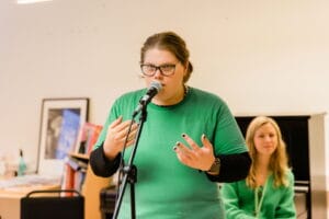 Person i grön tröja talar i mikrofon och gestikulerar, med en sittande person i bakgrunden.