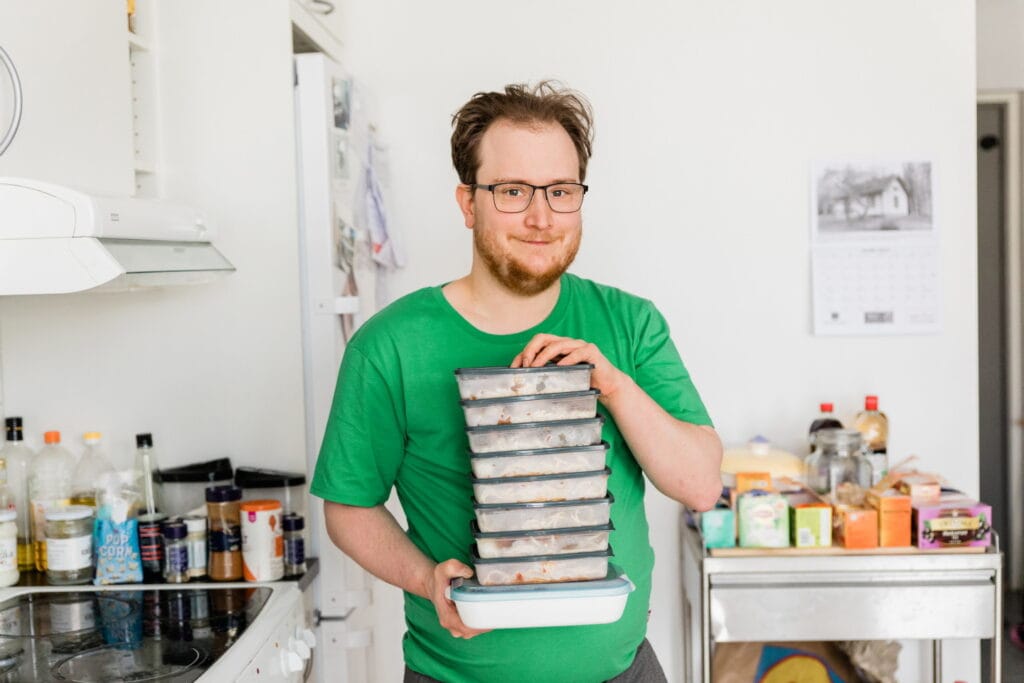 Man i gröna kläder står i ett kök och håller en stapel med matlådor fyllda med färdiglagad mat, med kryddor och livsmedel synliga i bakgrunden.