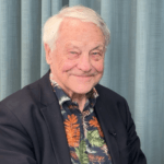 Äldre man i kavaj och mönstrad skjorta sitter framför blå gardin och ler mot kameran.