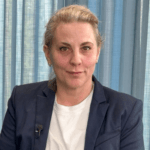 Kvinna i mörk kavaj och vit topp sitter framför blå gardin och tittar in i kameran.