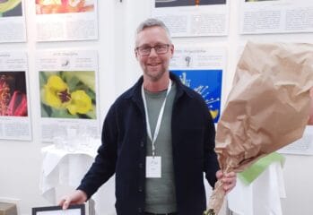 Man står och håller ett diplom och en stor bukett blommor, framför en vägg med blomsterbilder.