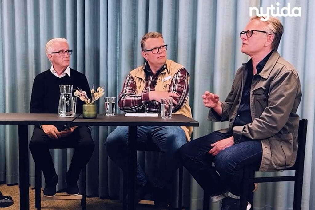 Tre personer sitter vid ett bord framför blå gardiner under ett samtal eller webbinarium. En person gestikulerar medan de två andra lyssnar. På bordet står en vattenkaraff, glas och en liten blomsterdekoration. Nytidas logotyp syns i bildens övre högra hörn.