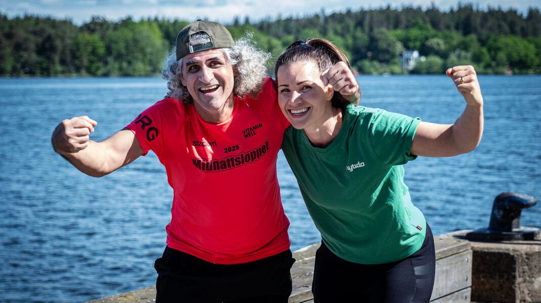 Salam och coach Caroline står vid vattnet, ler mot kameran och lyfter armarna i en peppande pose inför Glada Midnattsloppet.