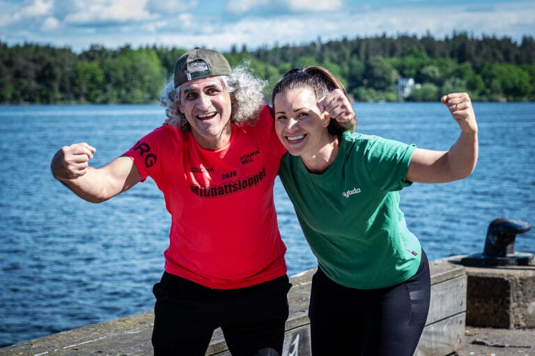 Salam och coach Caroline står vid vattnet, ler mot kameran och lyfter armarna i en peppande pose inför Glada Midnattsloppet.