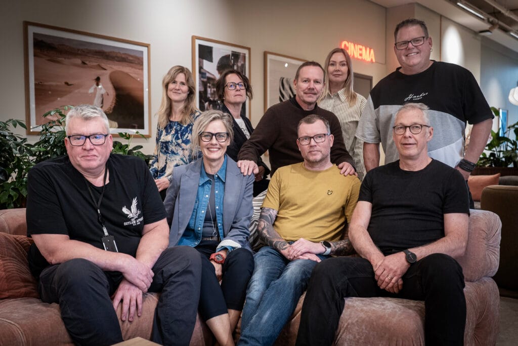 Gruppbild på nio personer samlade i en lounge med soffor och fåtöljer. Några sitter och andra står bakom, alla vända mot kameran. I bakgrunden syns växter, inramad konst på väggarna och en neonskylt med texten ”Cinema”, vilket skapar en varm och inbjudande miljö.