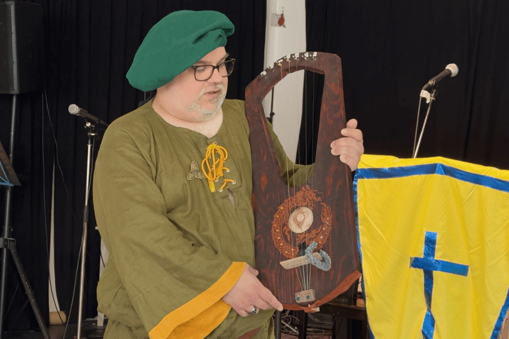 En person i grön medeltidsinspirerad dräkt och basker håller upp ett stränginstrument framför en scen med mikrofoner och svarta draperier. Bredvid syns en gul och blå sköld eller fana.