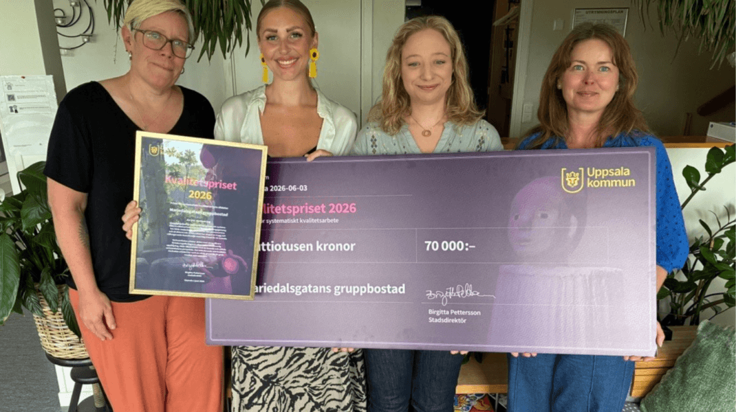 Fyra personer står inomhus och håller upp ett diplom och en stor prischeck från Uppsala kommun för Kvalitetspriset 2026. Checken är på 70 000 kronor och gäller Mariedalsgatans gruppbostad.
